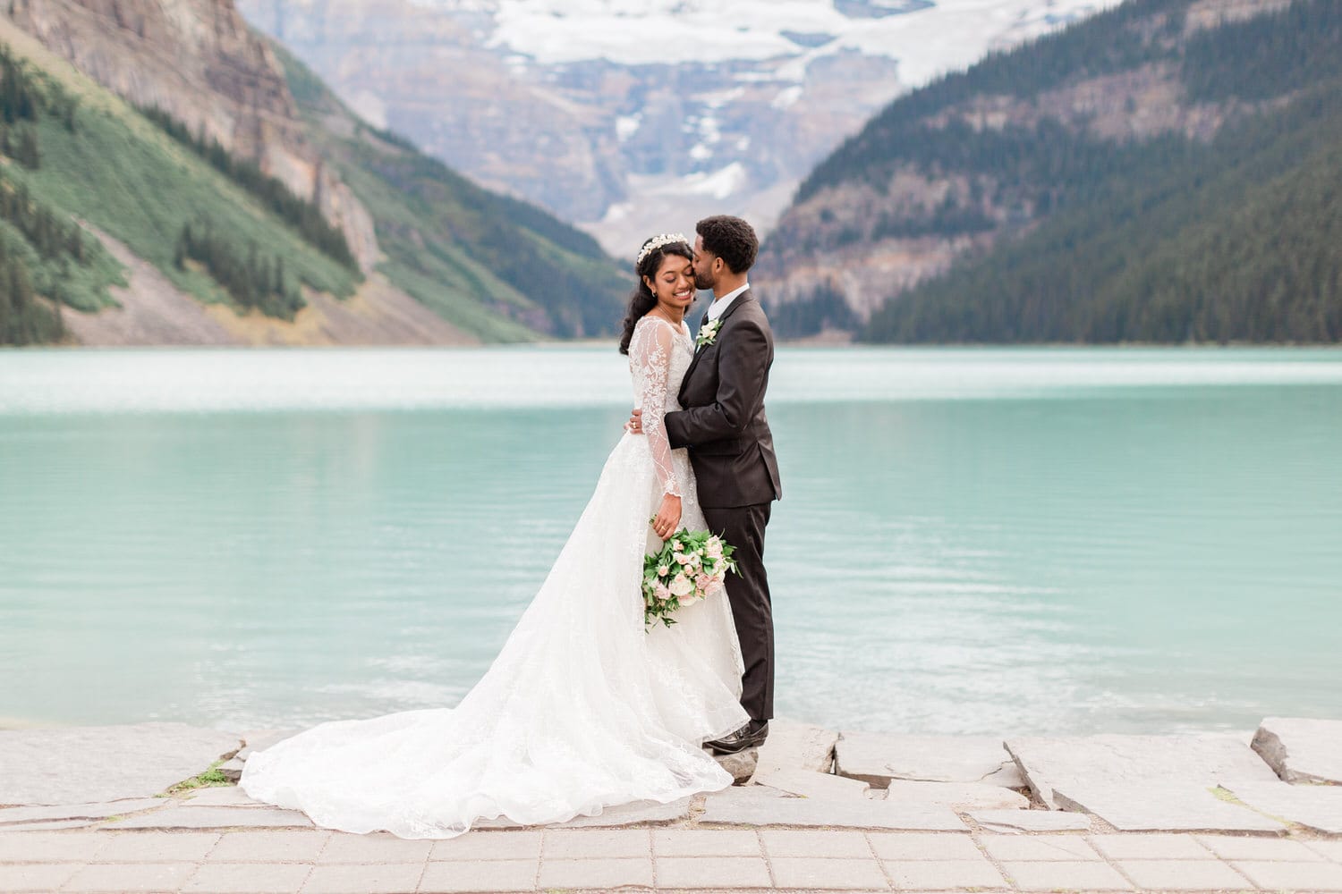 A couple in formal attire sharing a tender moment near a serene turquoise lake surrounded by mountains.