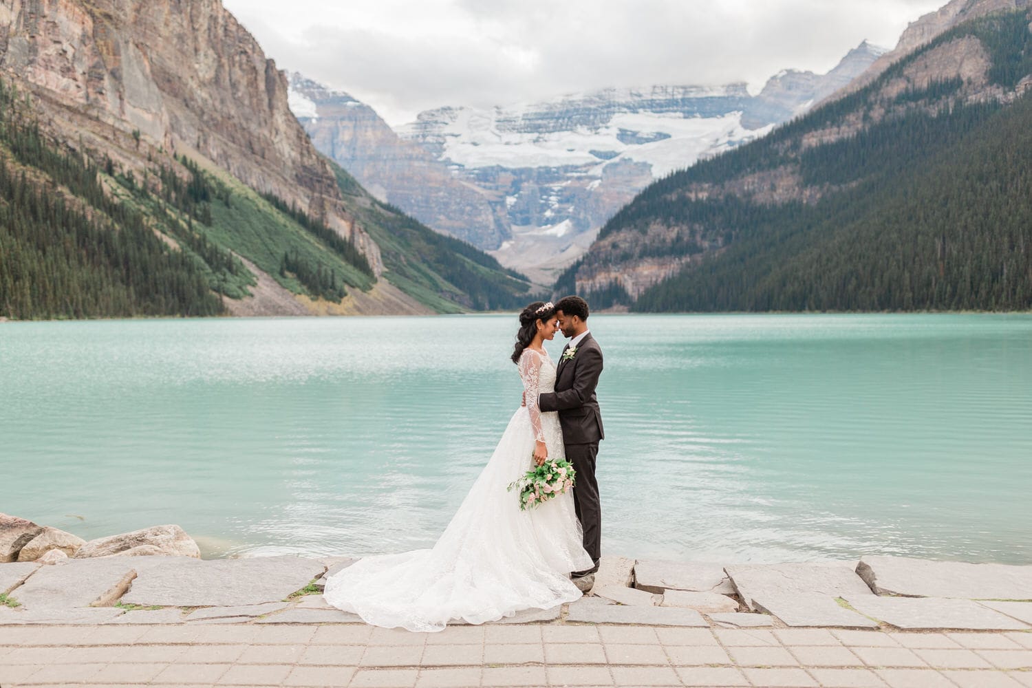 A bride and groom share a tender moment by a serene turquoise lake surrounded by majestic mountains and lush greenery.