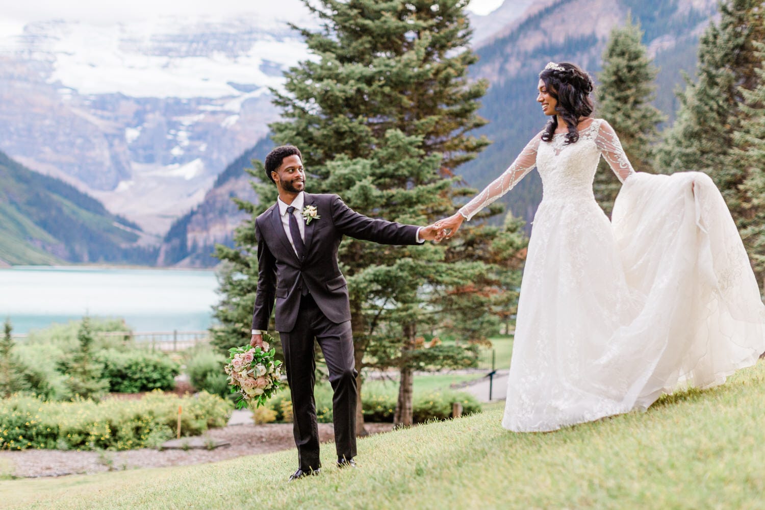 A couple joyfully holding hands in a picturesque outdoor setting with mountains and a lake in the background, showcasing the bride's elegant white gown and the groom's stylish suit.