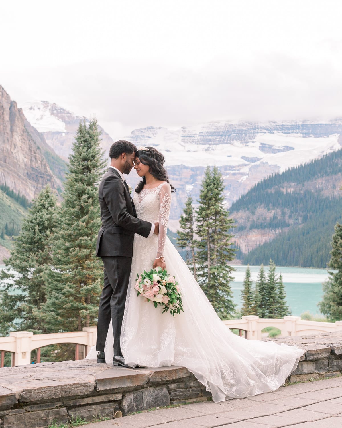 A couple sharing a tender moment with a stunning mountain and lake backdrop, the bride in a lace gown holding a bouquet of flowers.