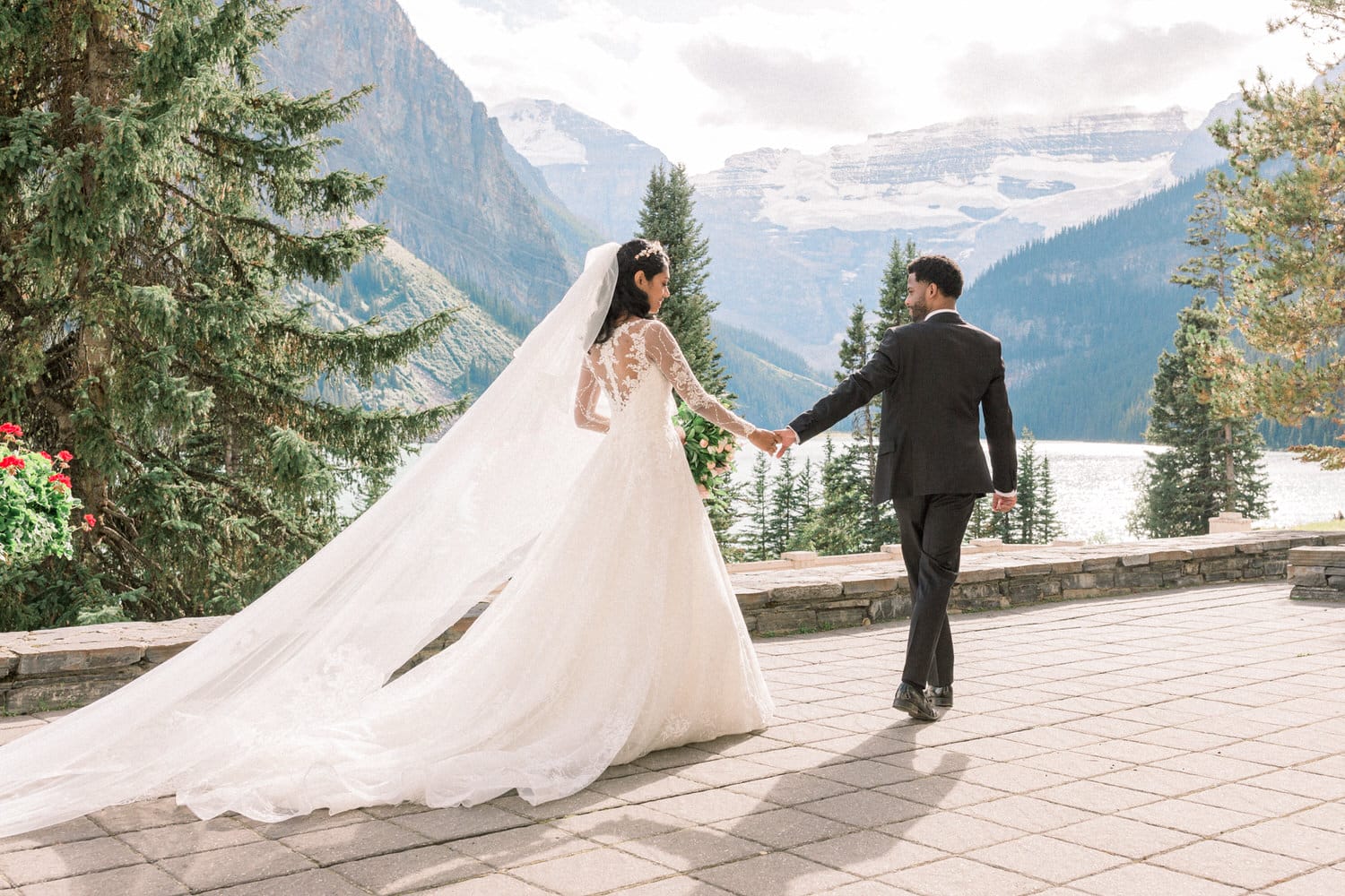 A bride and groom walking hand in hand against a stunning mountain backdrop, with lush greenery and a serene lake in the distance.