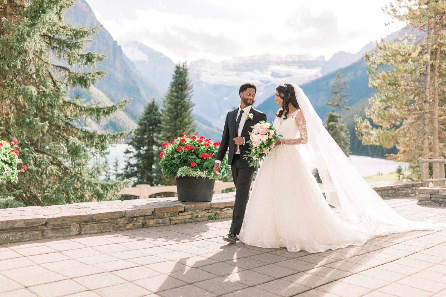 A joyful bride and groom walk hand in hand, surrounded by lush greenery and vibrant flowers, with mountains and a serene lake in the background.