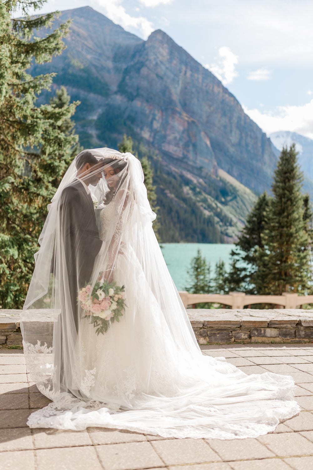 A couple sharing a tender moment under a veil with a scenic mountain backdrop and a serene lake.