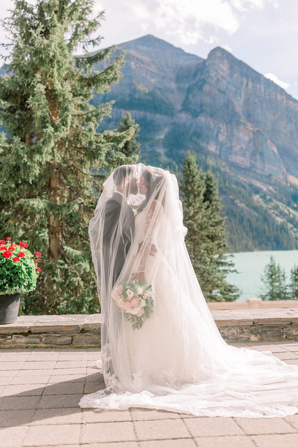 A bride and groom share a tender embrace under a sheer veil, framed by lush greenery and majestic mountains in the background.