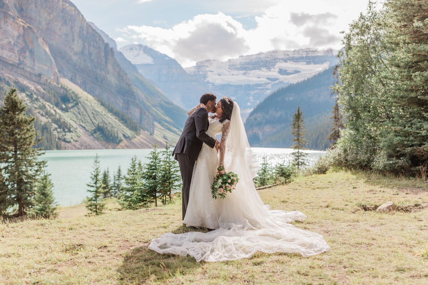 A couple sharing a kiss in wedding attire by a serene lake, surrounded by mountains and greenery.