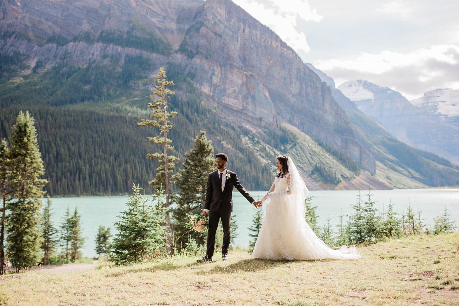 A bride and groom hold hands in a scenic landscape, surrounded by mountains and a lake, capturing a beautiful outdoor wedding moment.