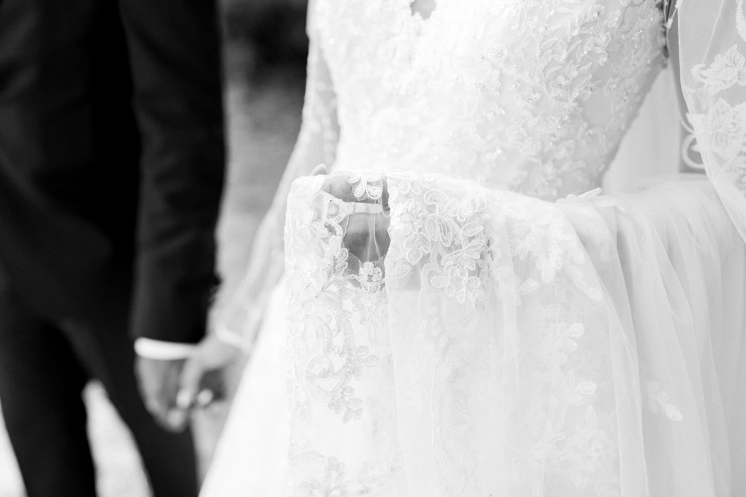 Close-up of a bridal gown with intricate lace detailing, paired with a glimpse of the groom's suit as they hold hands.