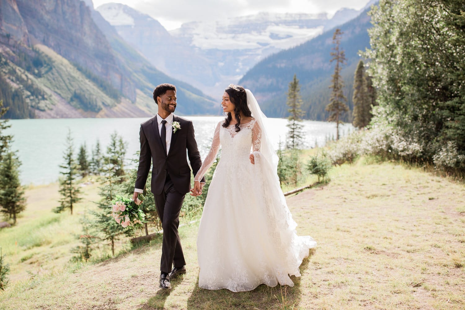 A joyful couple walks hand in hand by a serene lake, surrounded by lush greenery and mountains.