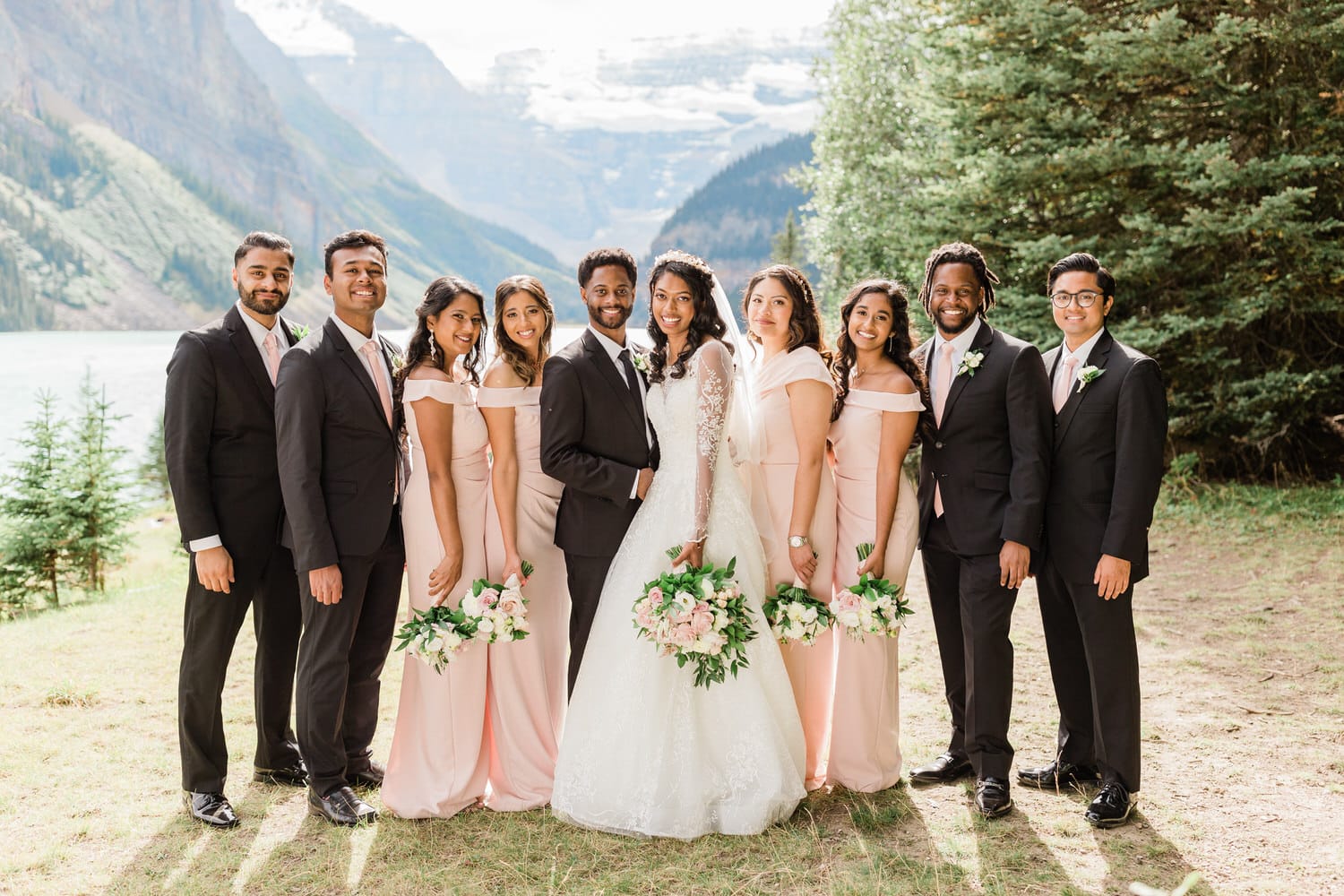 A joyful bridal party poses outdoors, featuring the bride in a stunning white gown surrounded by bridesmaids in elegant pink dresses, with a picturesque mountain backdrop and lake.