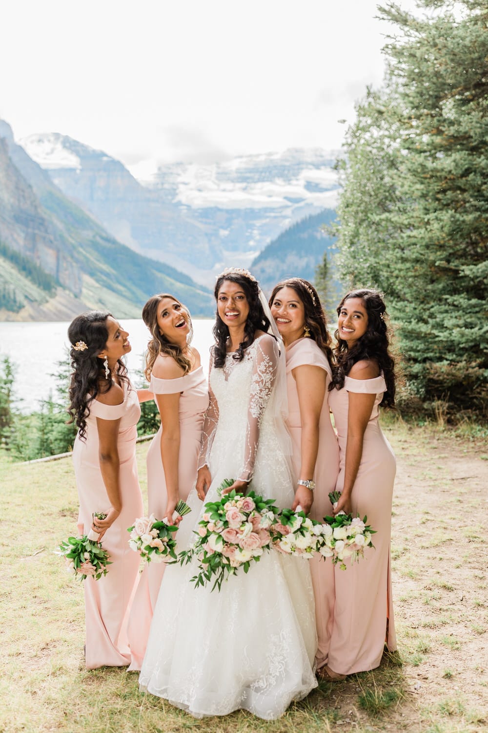 A joyful bride in a white gown surrounded by her bridesmaids in soft pink dresses, all smiling in a breathtaking natural setting with mountains and a lake in the background.