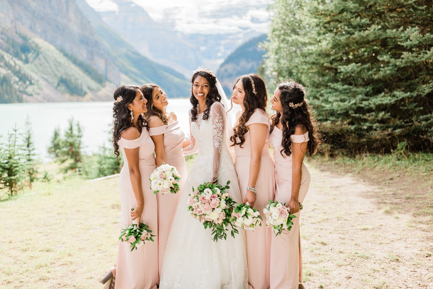 A joyful bride standing with her five bridesmaids in elegant pink dresses, surrounded by a scenic backdrop of mountains and a serene lake. Each bridesmaid holds a bouquet of flowers, capturing a moment of happiness and camaraderie.