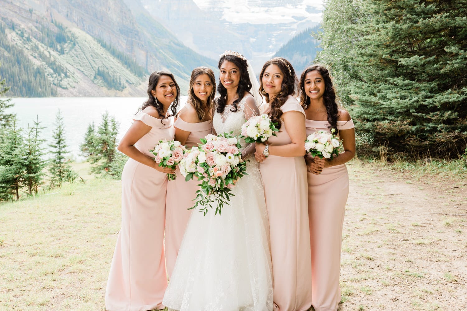 A joyful bride and her five bridesmaids pose together outdoors, surrounded by lush greenery and a scenic lake backdrop, all wearing matching pink dresses and holding beautiful floral bouquets.