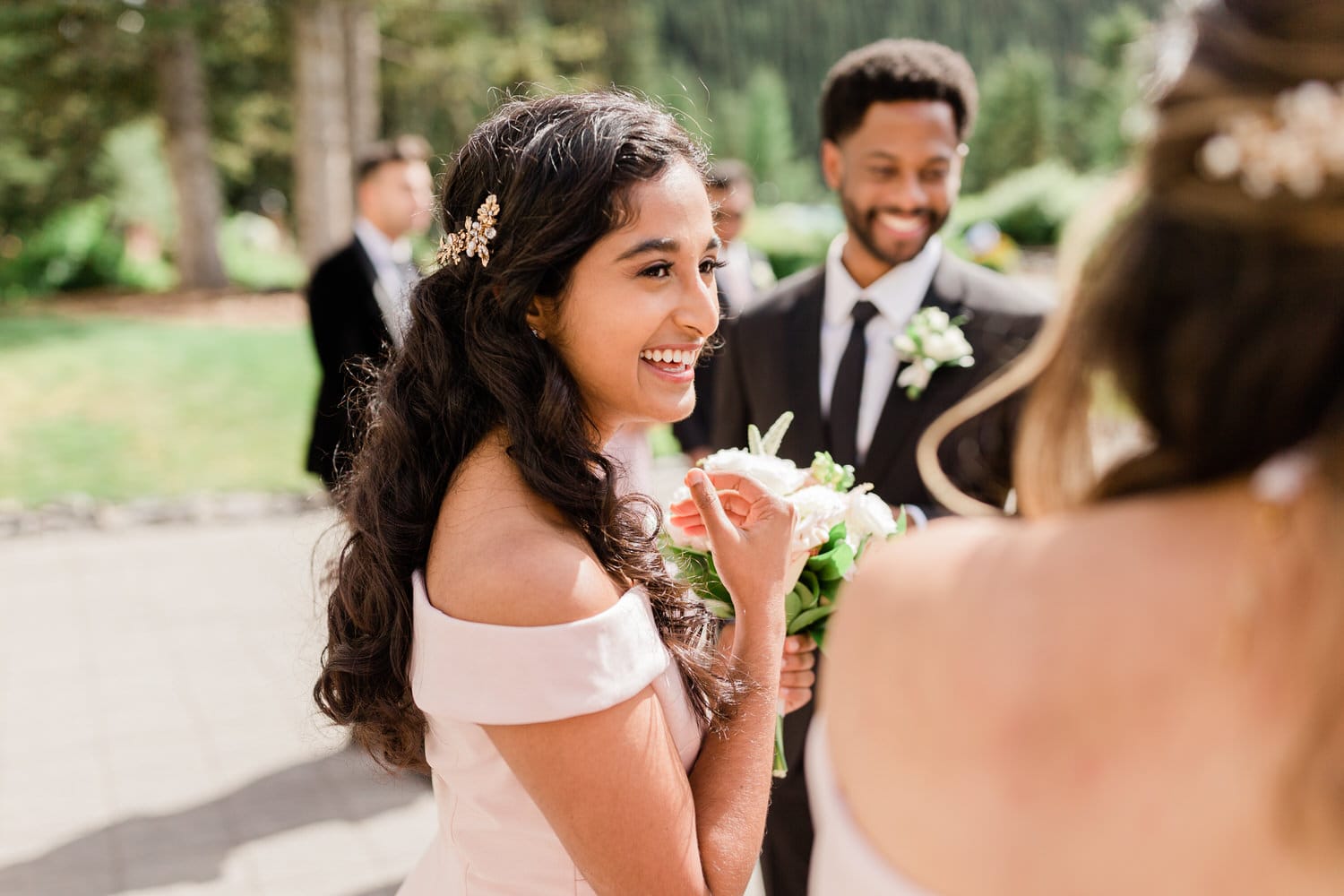 A young woman in an off-shoulder pink dress smiles brightly while holding a bouquet, surrounded by friends in formal attire, with lush greenery in the background.