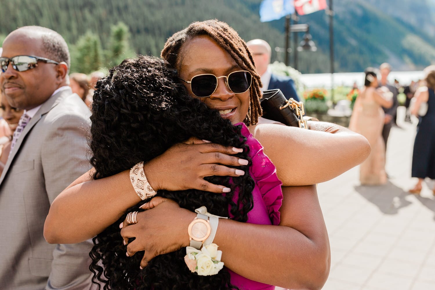 Two women embrace warmly in a heartfelt hug during an outdoor event, with a man in sunglasses visible in the background.