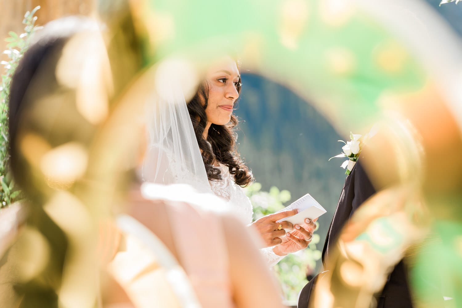 A bride holding a wedding vow card, captured through a blurred ornamental frame, during an outdoor ceremony with lush greenery and mountains in the background.