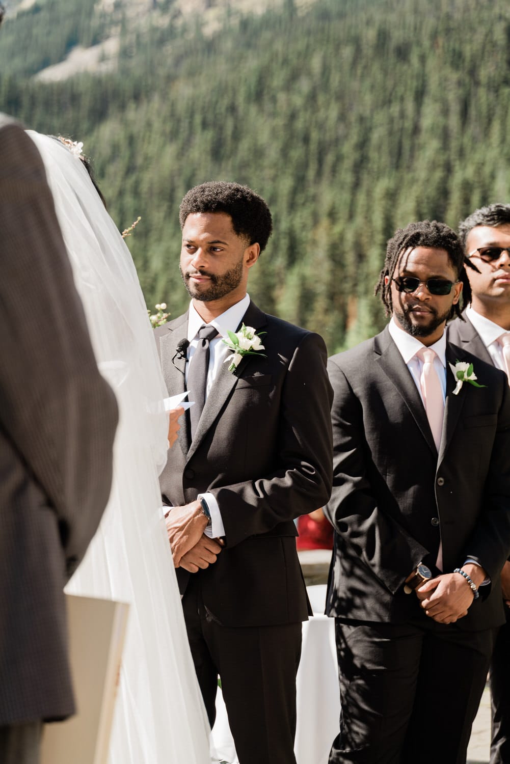 Groom and groomsmen standing attentively during an outdoor wedding ceremony, surrounded by lush greenery.