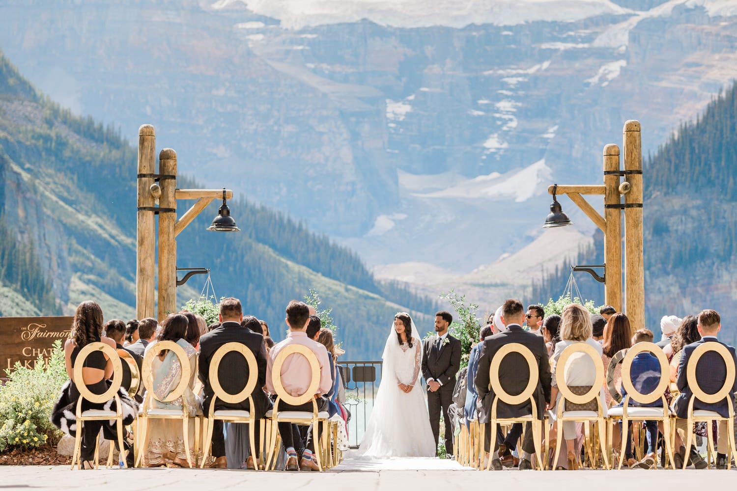 A bride and groom stand at the altar with guests seated in elegant chairs, surrounded by a stunning mountain backdrop during a wedding ceremony.