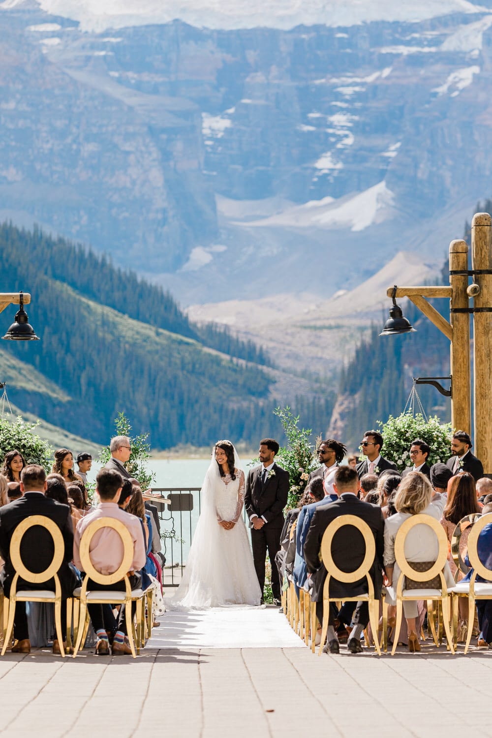 A bride and groom stand together at the altar during their wedding ceremony, surrounded by guests seated in elegant chairs with a stunning mountain landscape in the background.