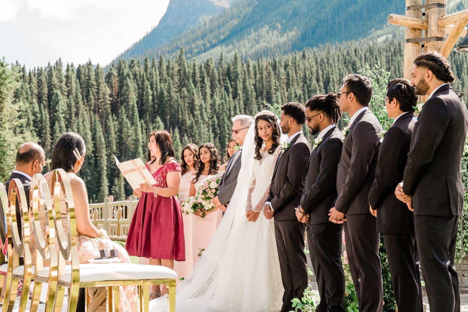 A bride and groom stand before an officiant, surrounded by wedding guests in a scenic outdoor setting with lush pine trees and mountains in the background.