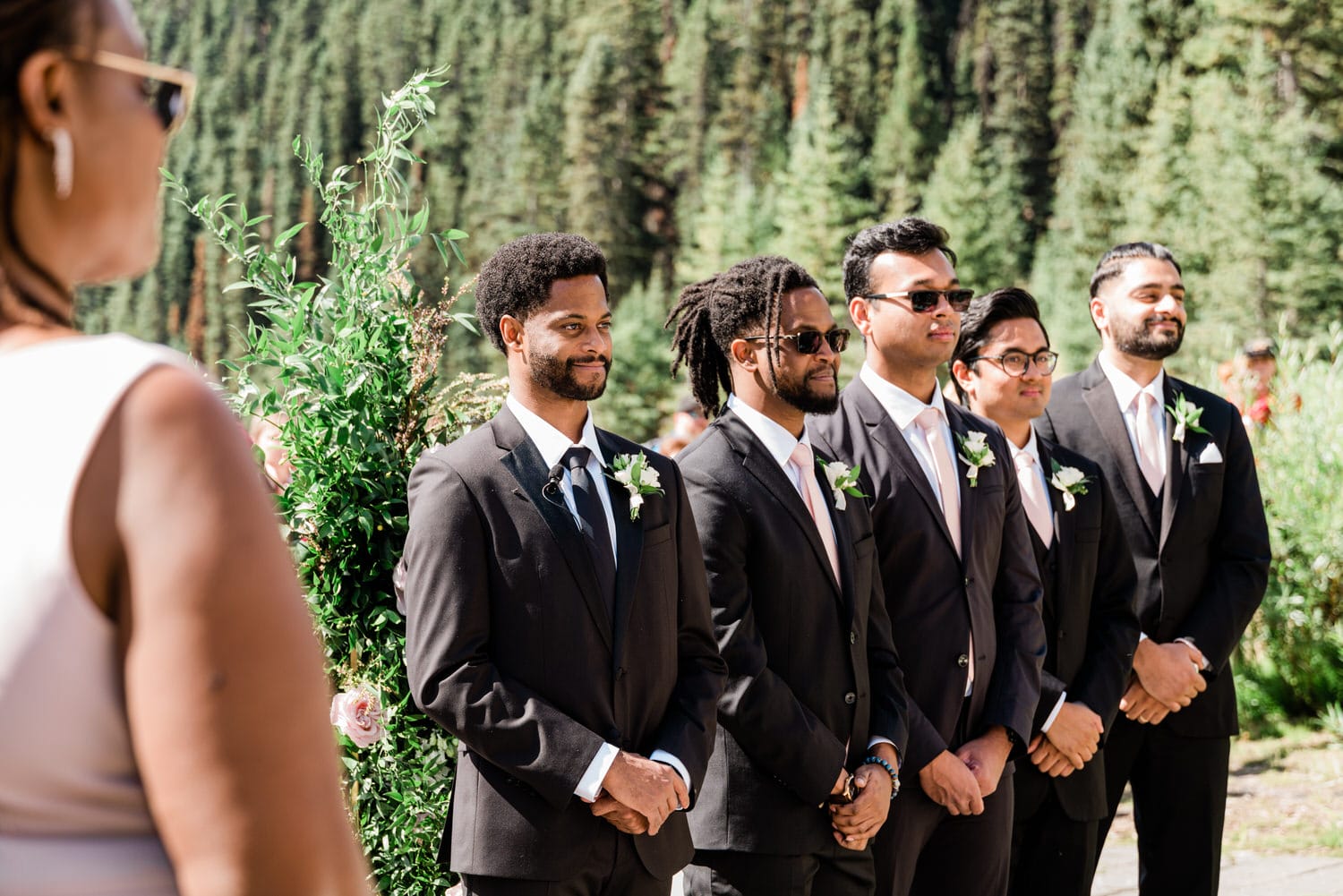 A group of well-dressed men standing in a wedding ceremony setting, surrounded by greenery and trees, with one woman partially visible in the foreground.