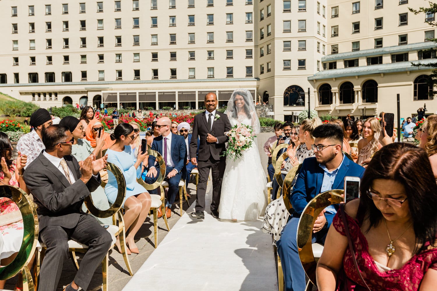 A bride escorted by her father walks down the aisle during a sunny outdoor wedding ceremony, surrounded by seated guests enjoying the moment.