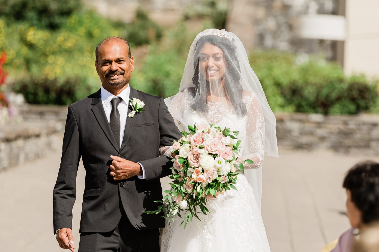 A smiling bride in a lace gown and veil walks down the aisle, escorted by a man in a suit, both radiating joy amidst a beautiful outdoor setting.