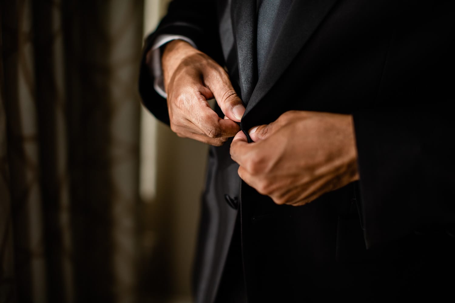 A close-up of a man adjusting the button of his black suit jacket, showcasing elegant attire and groomed hands.