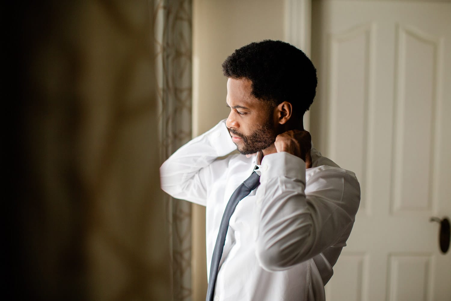 A man adjusting his tie while preparing for a formal event, captured in a softly lit room.