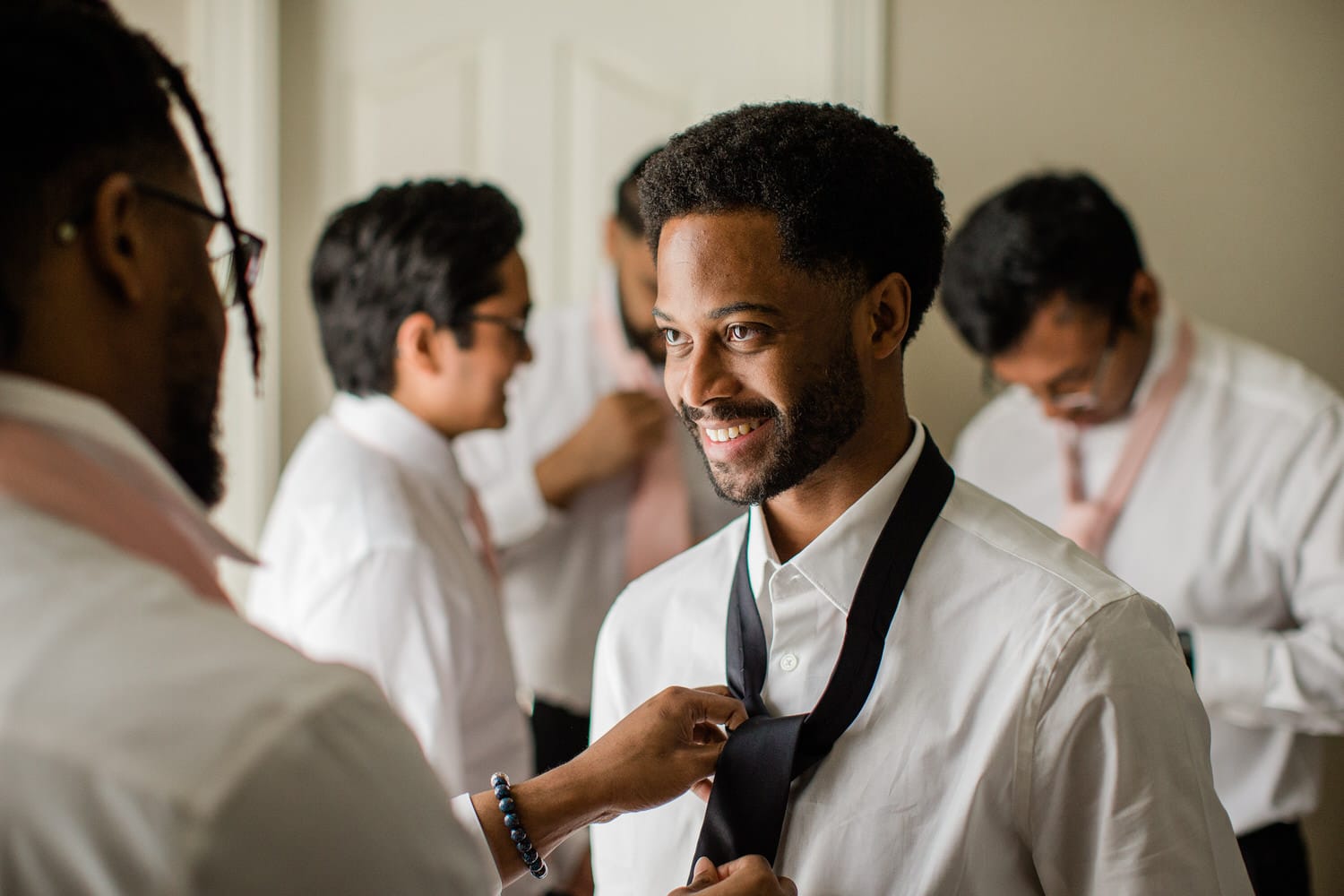 A group of well-dressed men, including the groom, smiles while adjusting ties in a bright room, showcasing camaraderie and celebration before the event.