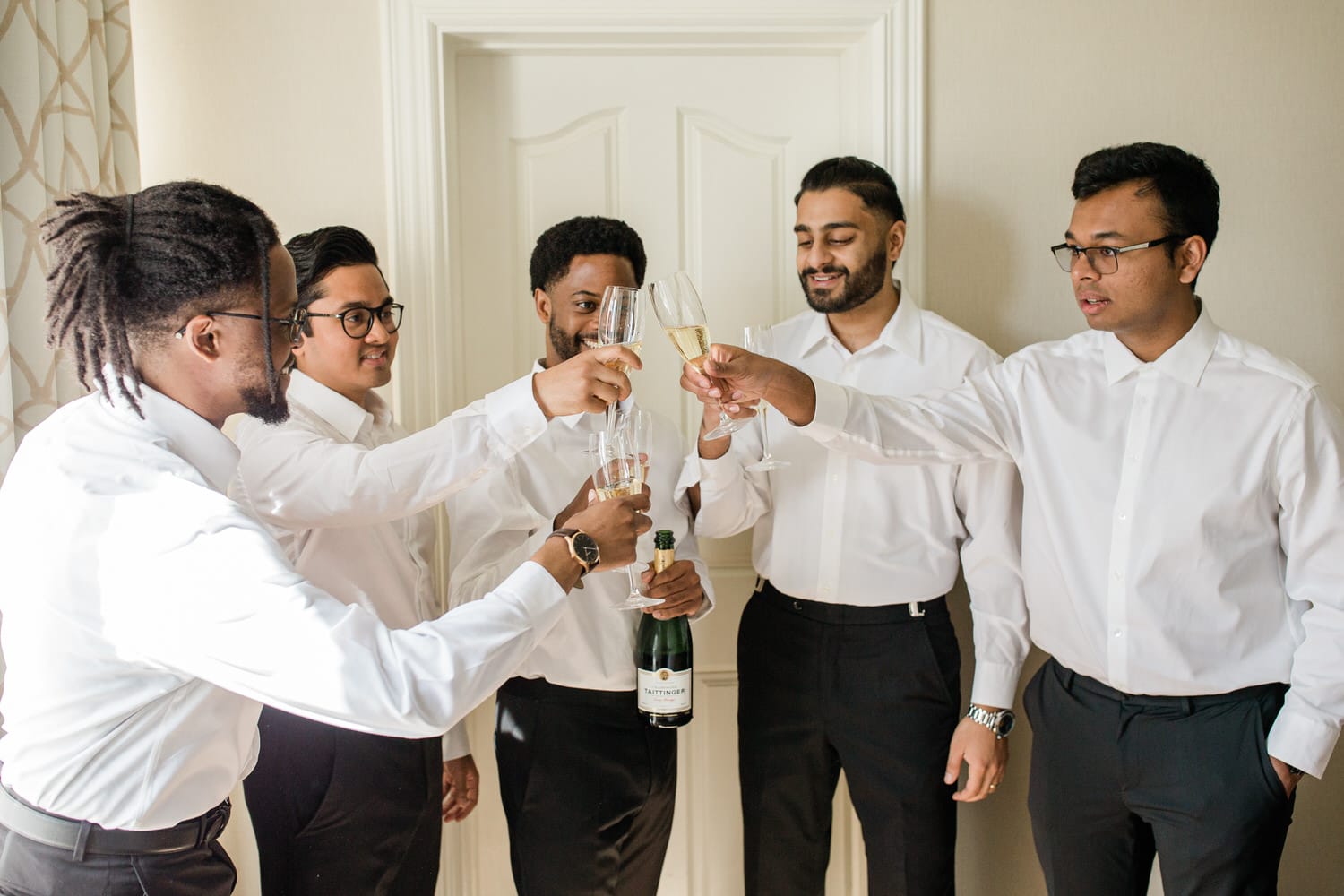 A group of five men in white shirts raising glasses in celebration, with a bottle of champagne in the center.