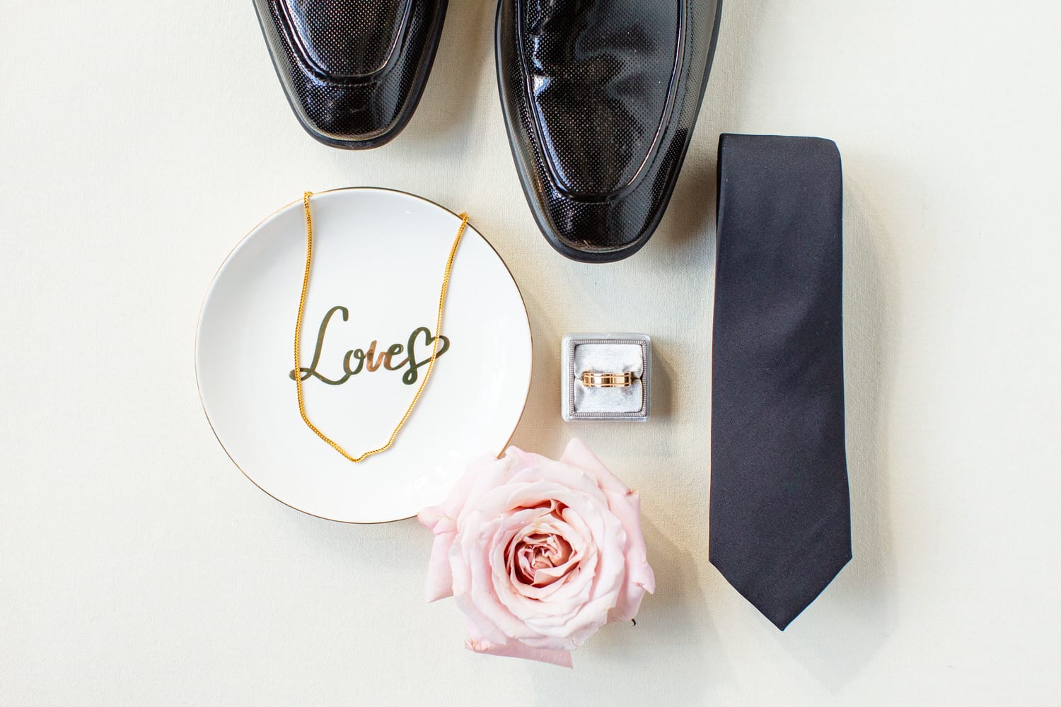 A flat lay featuring black formal shoes, a gold necklace with the word "Love," a silver ring in a box, a black tie, and a pink rose on a neutral background.