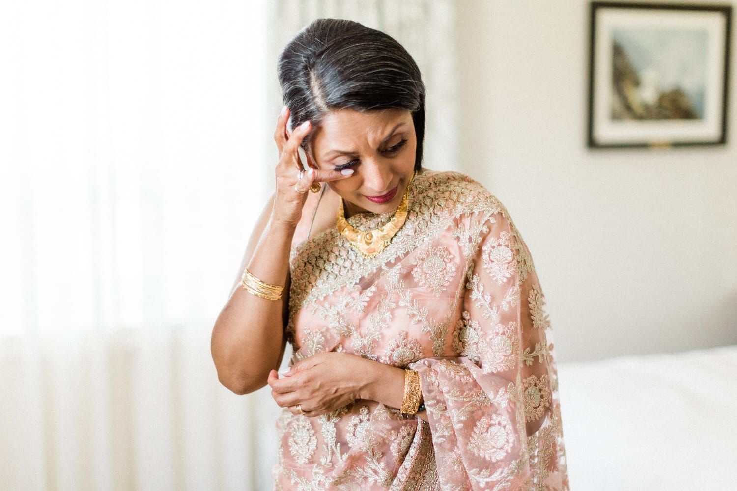 A woman wearing a beautifully embroidered pink sari, adorned with gold jewelry, appears contemplative as she gently touches her head, with soft natural light illuminating the scene.