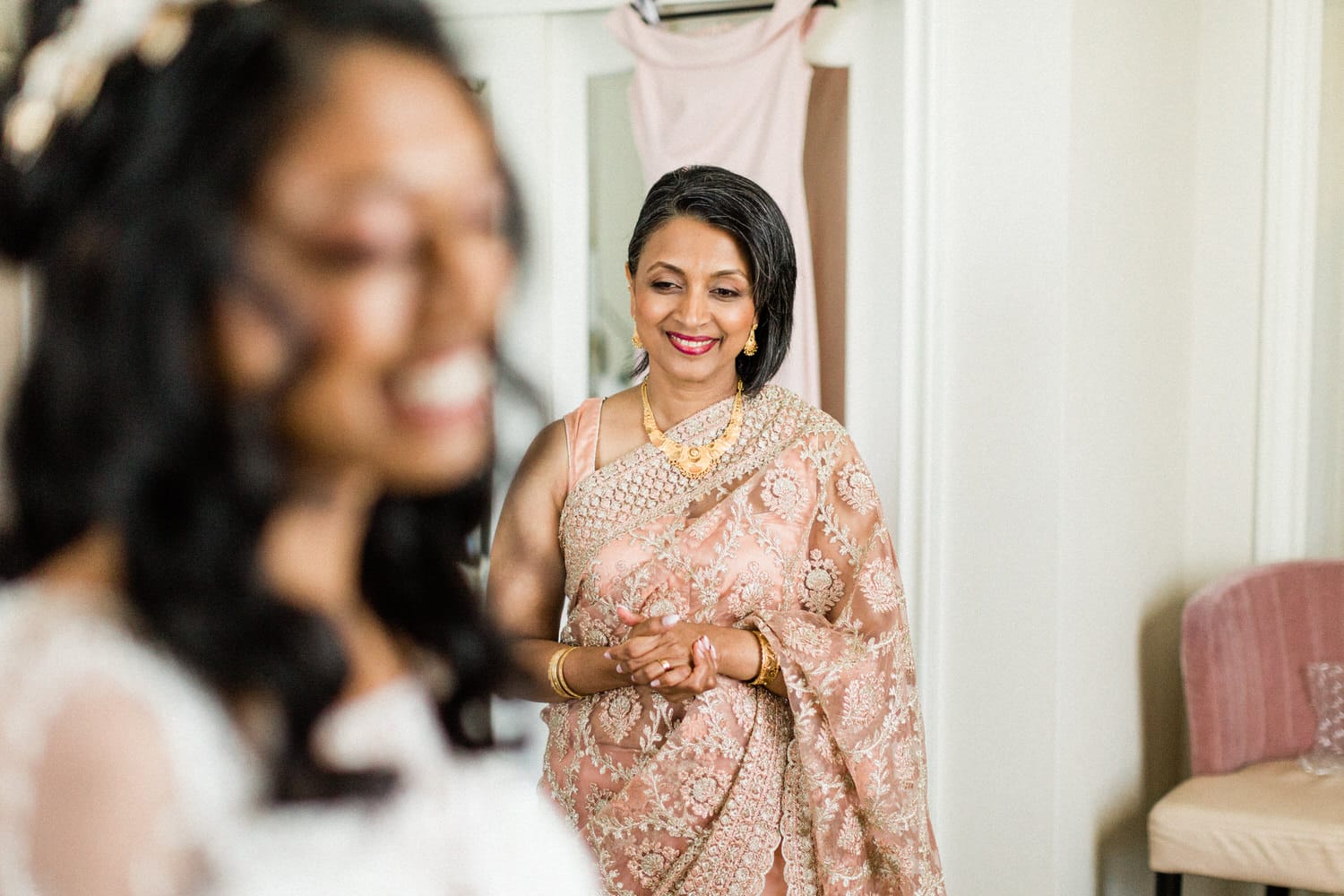 A mother in a beautiful saree smiles warmly as she watches her daughter getting ready, showcasing a moment of love and celebration.