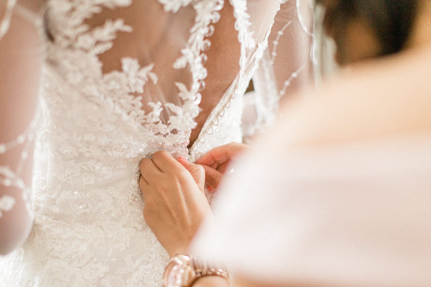 Close-up of a bride having her wedding dress buttoned by a helper, showcasing intricate lace detailing.