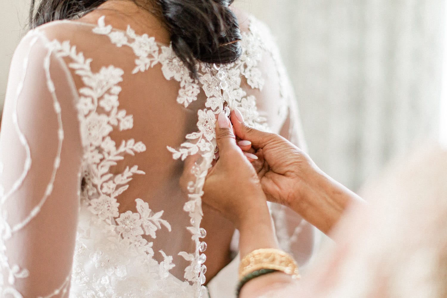 A close-up of a bride's back adorned with lace details as someone fastens the buttons of her wedding dress.