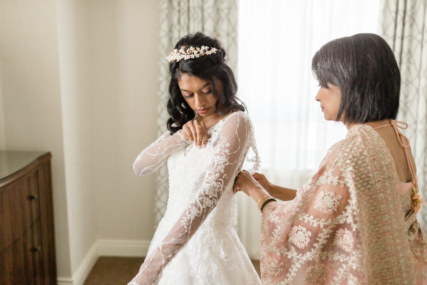 A bride adjusting her wedding dress while her mother assists her, surrounded by soft natural light in a cozy room.