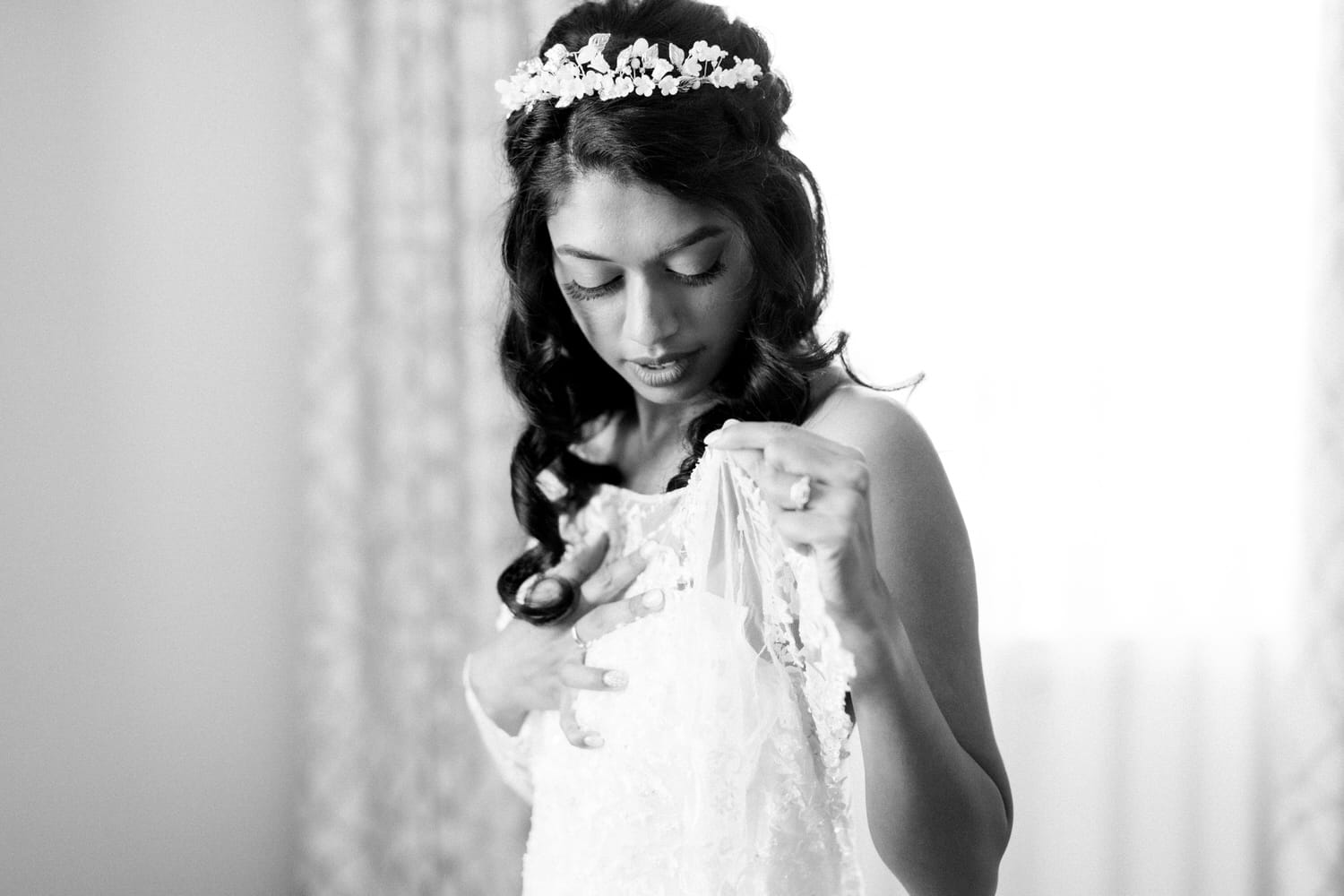 A bride in a delicate lace gown examines her dress, illuminated by natural light, showcasing her intricate hairstyle adorned with flowers.