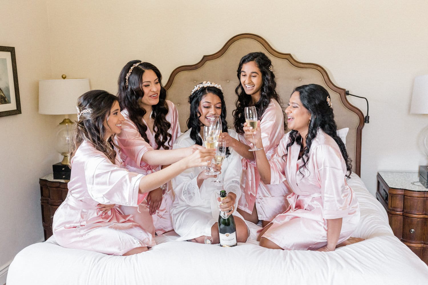 A joyful bridal party in pink robes toasting with glasses of champagne on a bed, capturing a moment of happiness and camaraderie before the wedding.