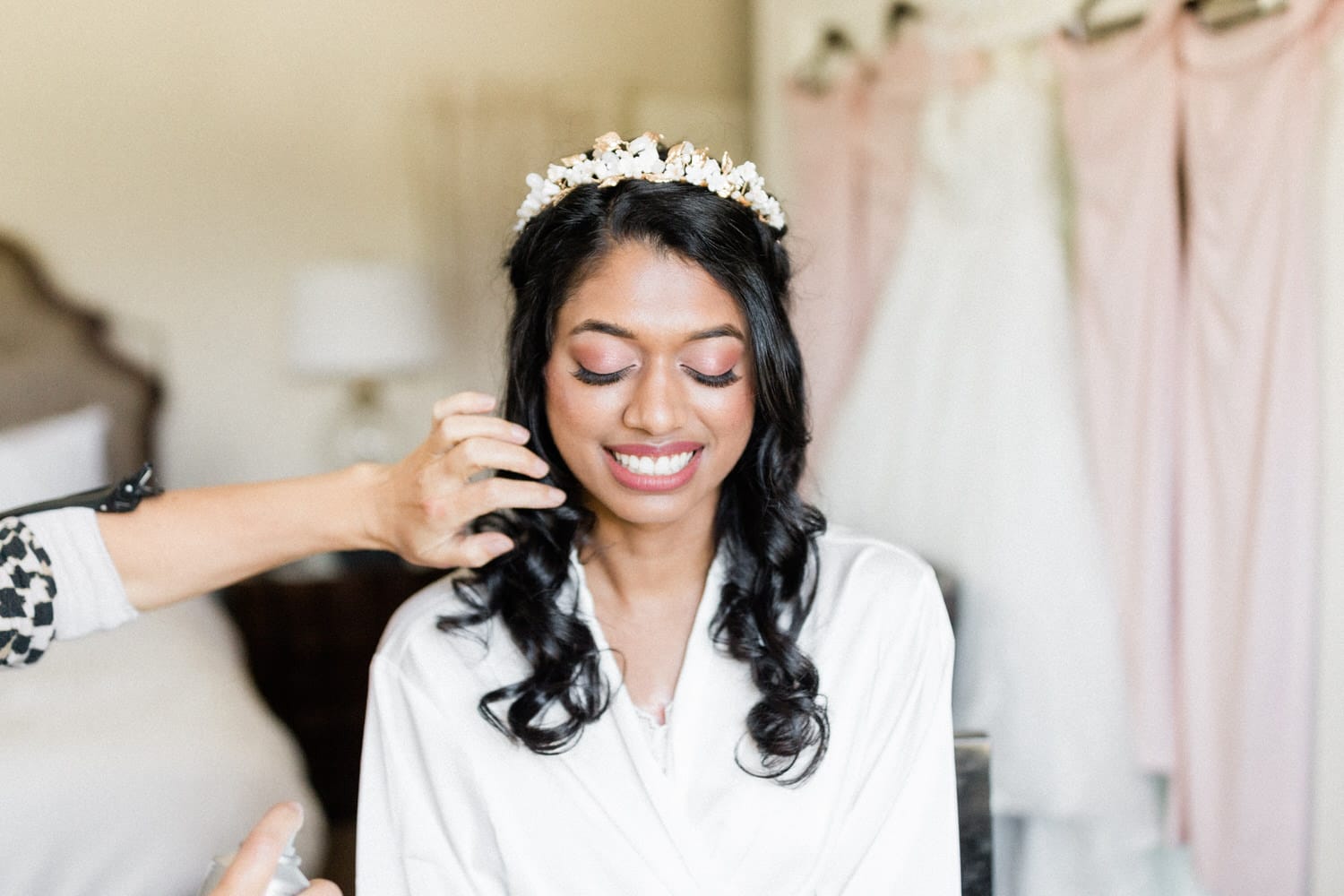 A bride getting her hair styled while wearing a robe, smiling with joy, in a softly lit room.