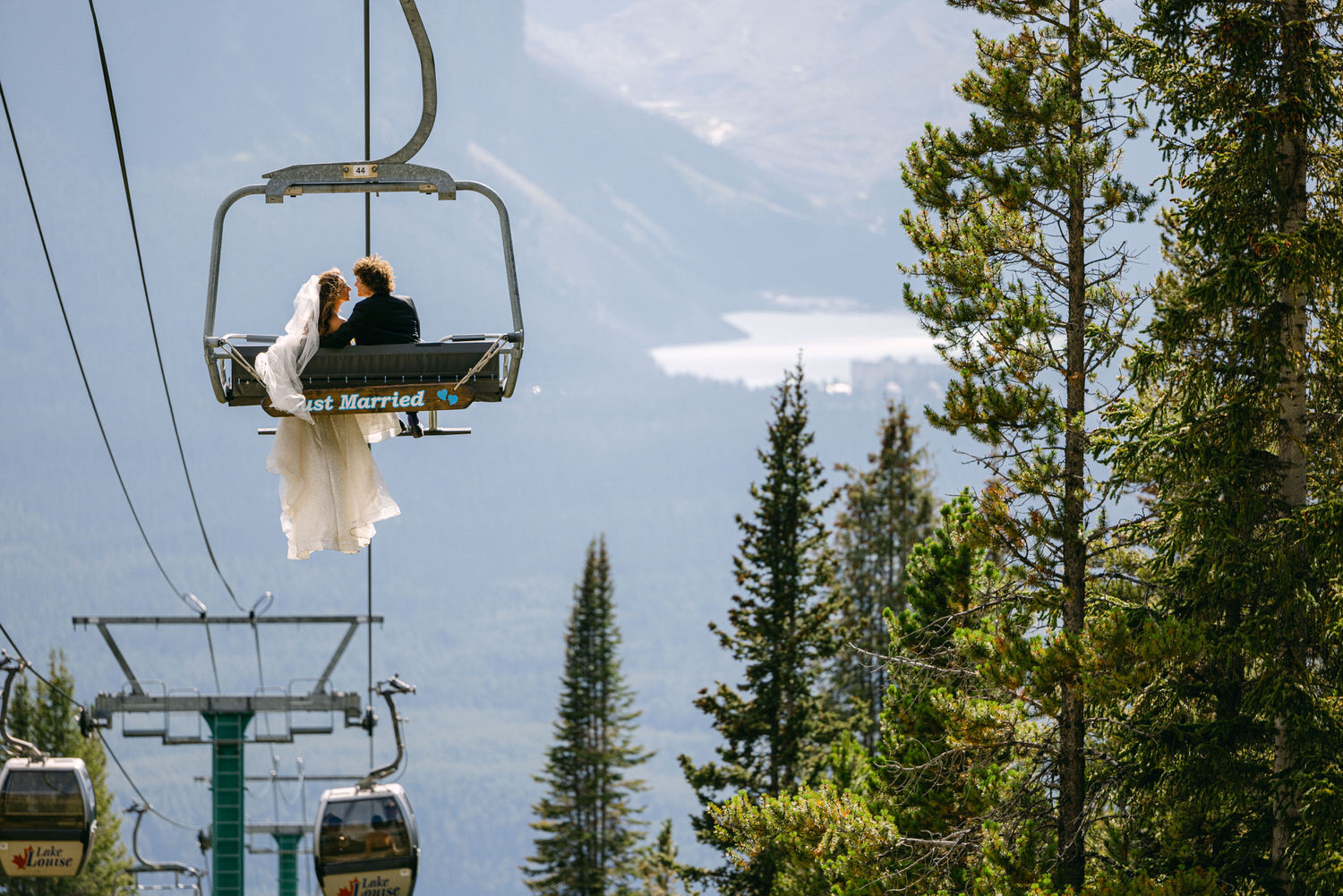 A newlywed couple shares a moment of joy while riding a chairlift, surrounded by picturesque mountains and trees, with a sign reading "Just Married."