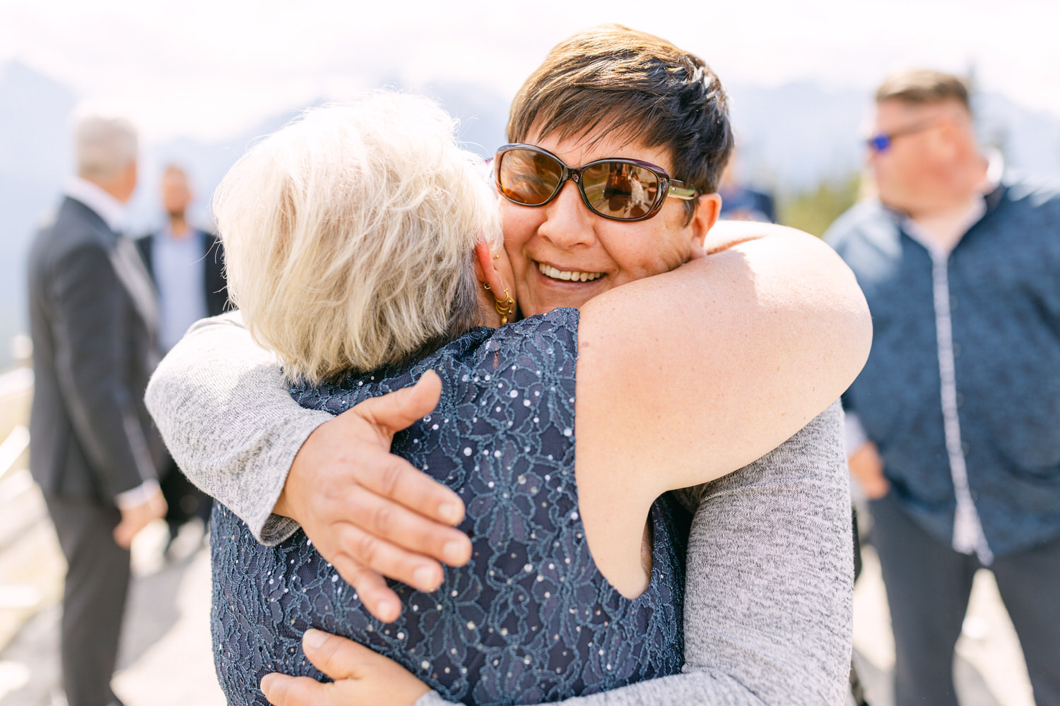 Two women embrace warmly, smiling in a beautiful outdoor setting with mountains in the background, capturing a moment of happiness and connection.