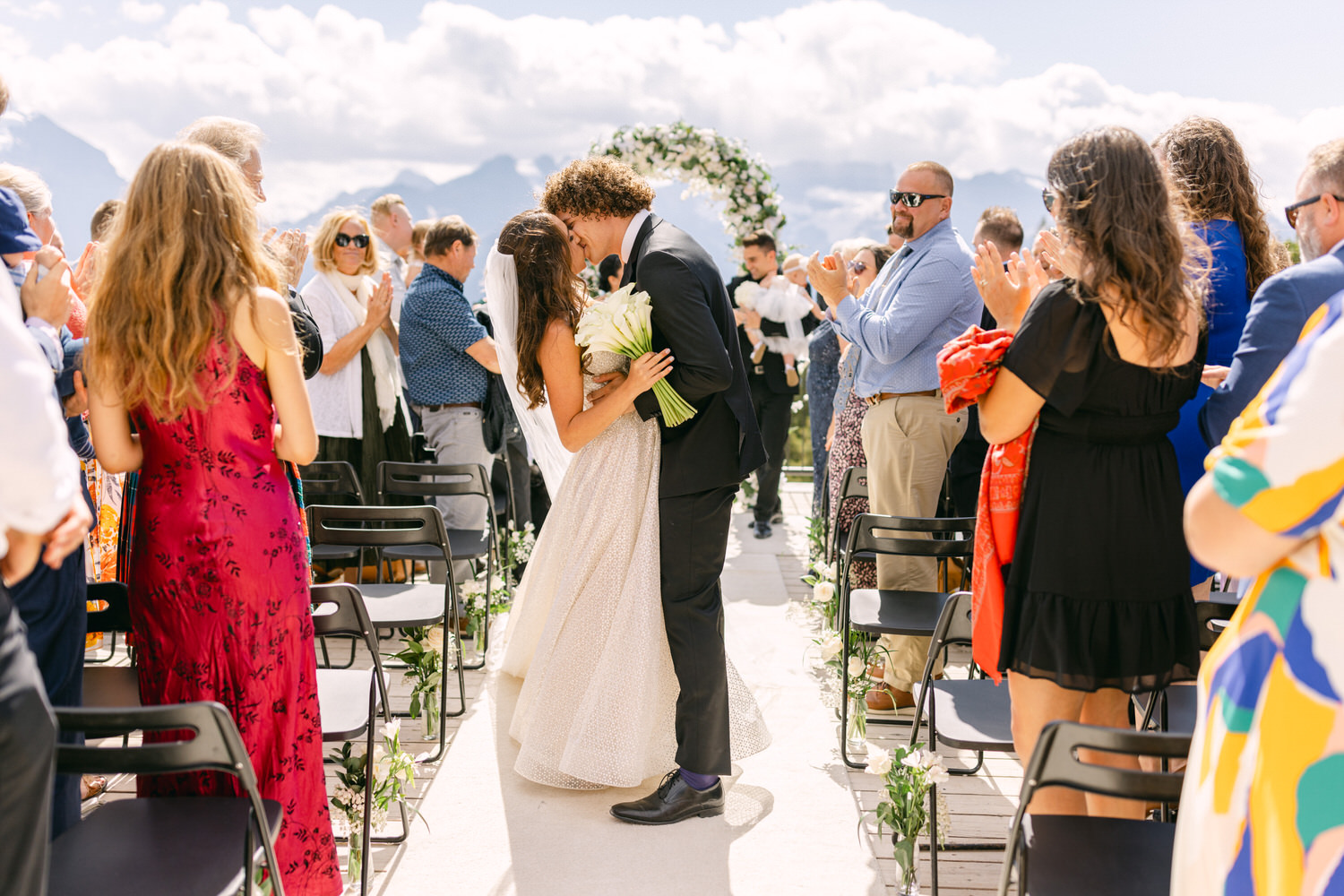 A bride and groom share a kiss during their outdoor wedding ceremony, surrounded by applauding guests and a picturesque mountain backdrop.