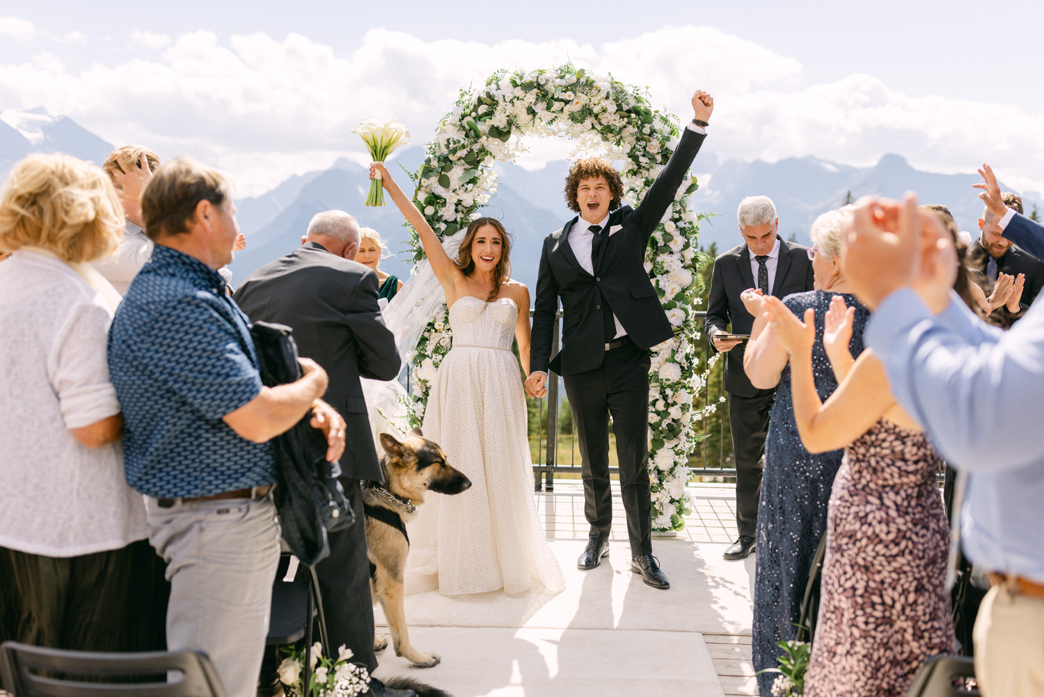 A joyful couple celebrates their marriage under a floral arch, surrounded by guests and a dog amidst a scenic mountain backdrop.