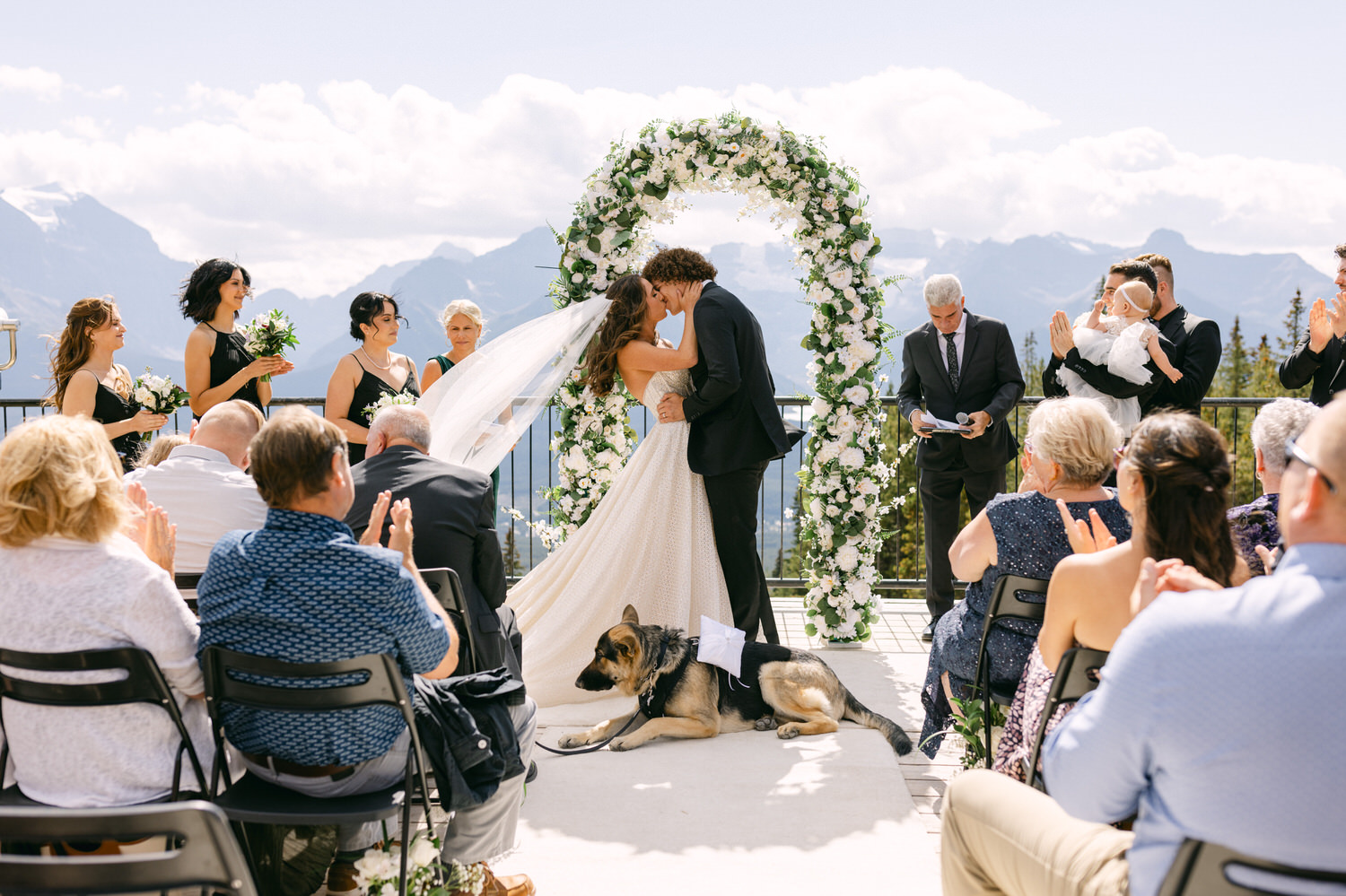 Couple sharing a kiss under a floral arch with guests and a dog in attendance at a scenic outdoor wedding.