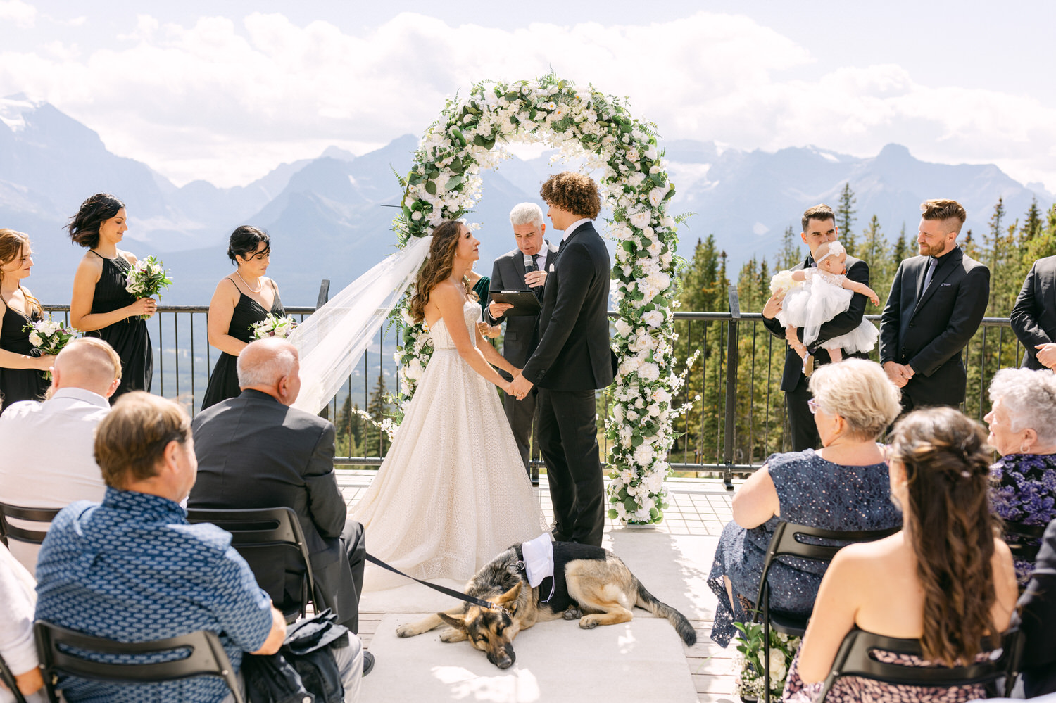 A beautiful outdoor wedding ceremony set in the mountains, featuring a couple exchanging vows under a floral arch, surrounded by guests and a dog in a tuxedo.