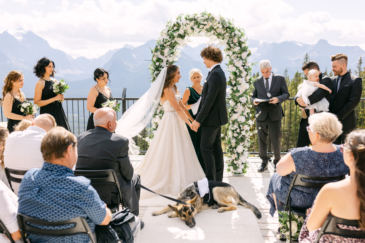 A bride and groom exchange vows at an outdoor ceremony adorned with floral decorations and surrounded by guests, including a dog lounging at their feet.