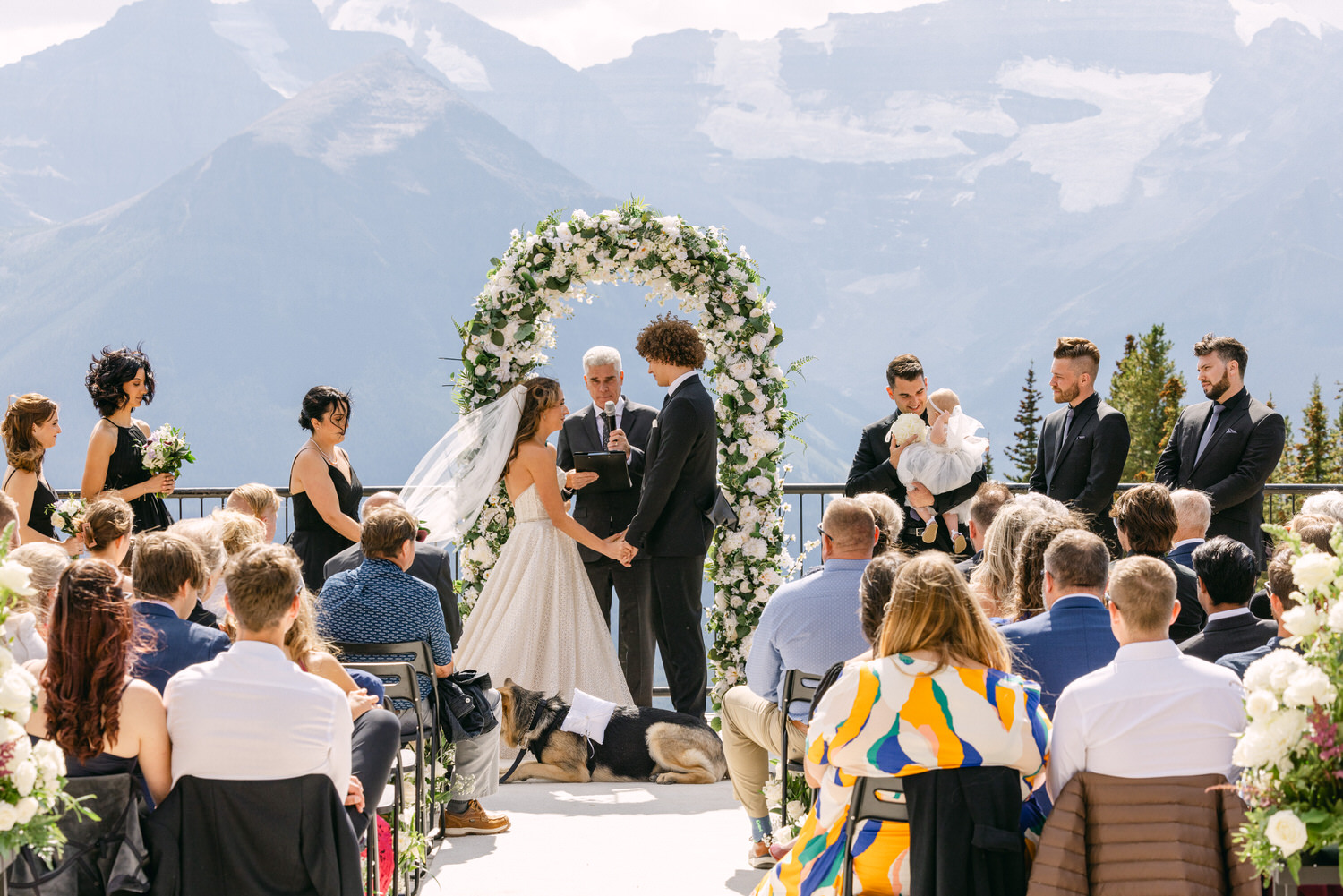 A couple exchanges vows under a floral arch with stunning mountain scenery, surrounded by guests and bridal party in a picturesque outdoor setting.