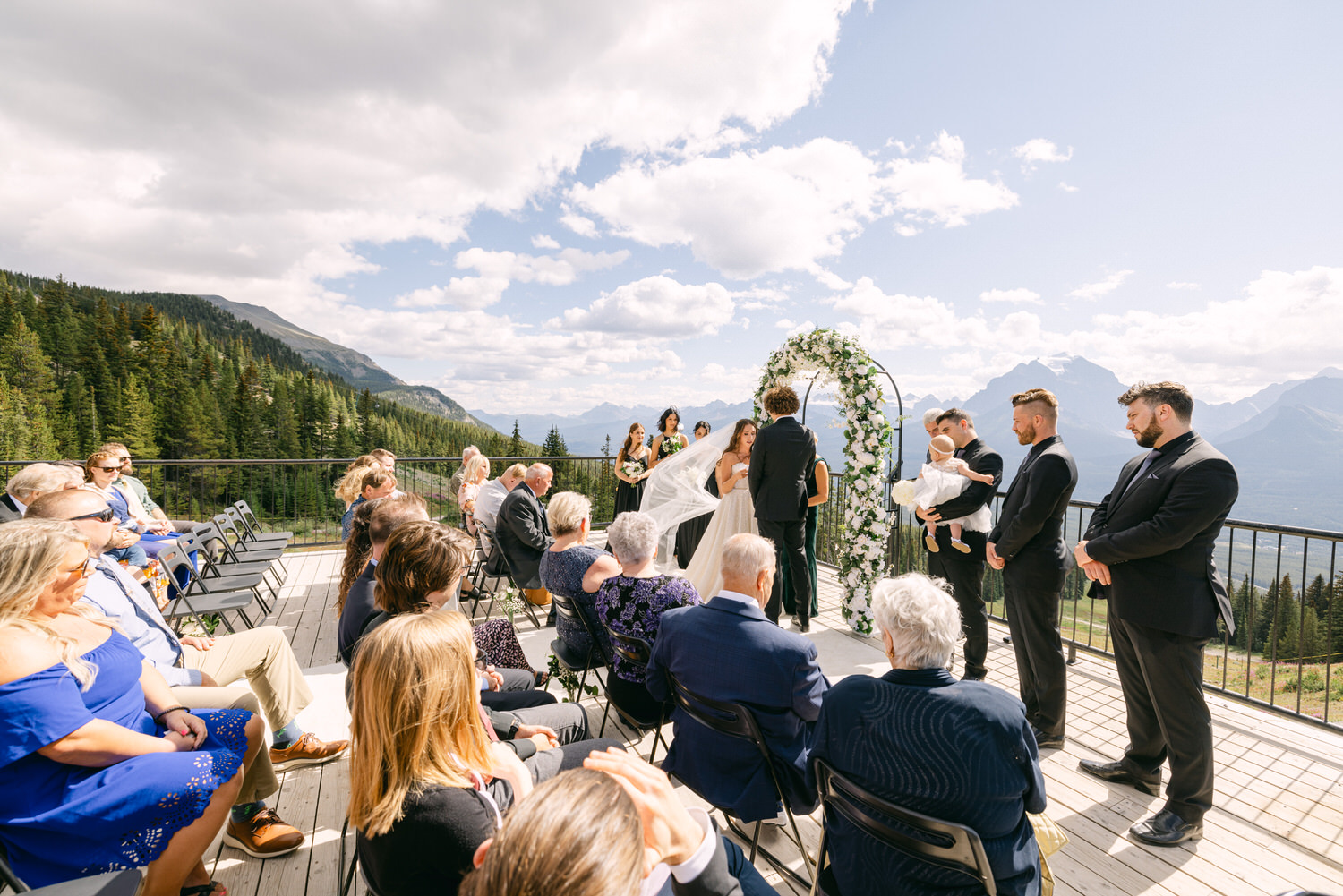 A couple exchanging vows under a floral arch on a balcony surrounded by guests and lush forests, with mountains in the backdrop.