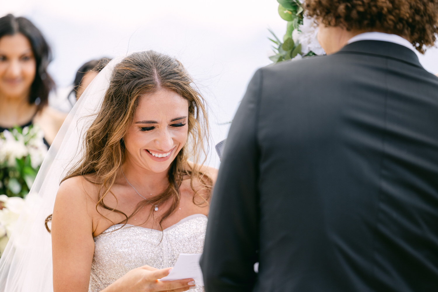 Bride smiling and reading vows during an outdoor wedding ceremony with wedding party in the background.