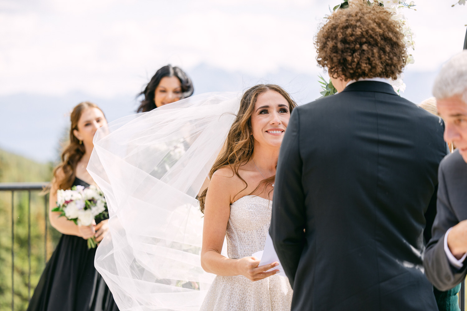 A joyful bride shares a moment with her groom during an outdoor wedding ceremony, surrounded by bridesmaids and beautiful mountain scenery.